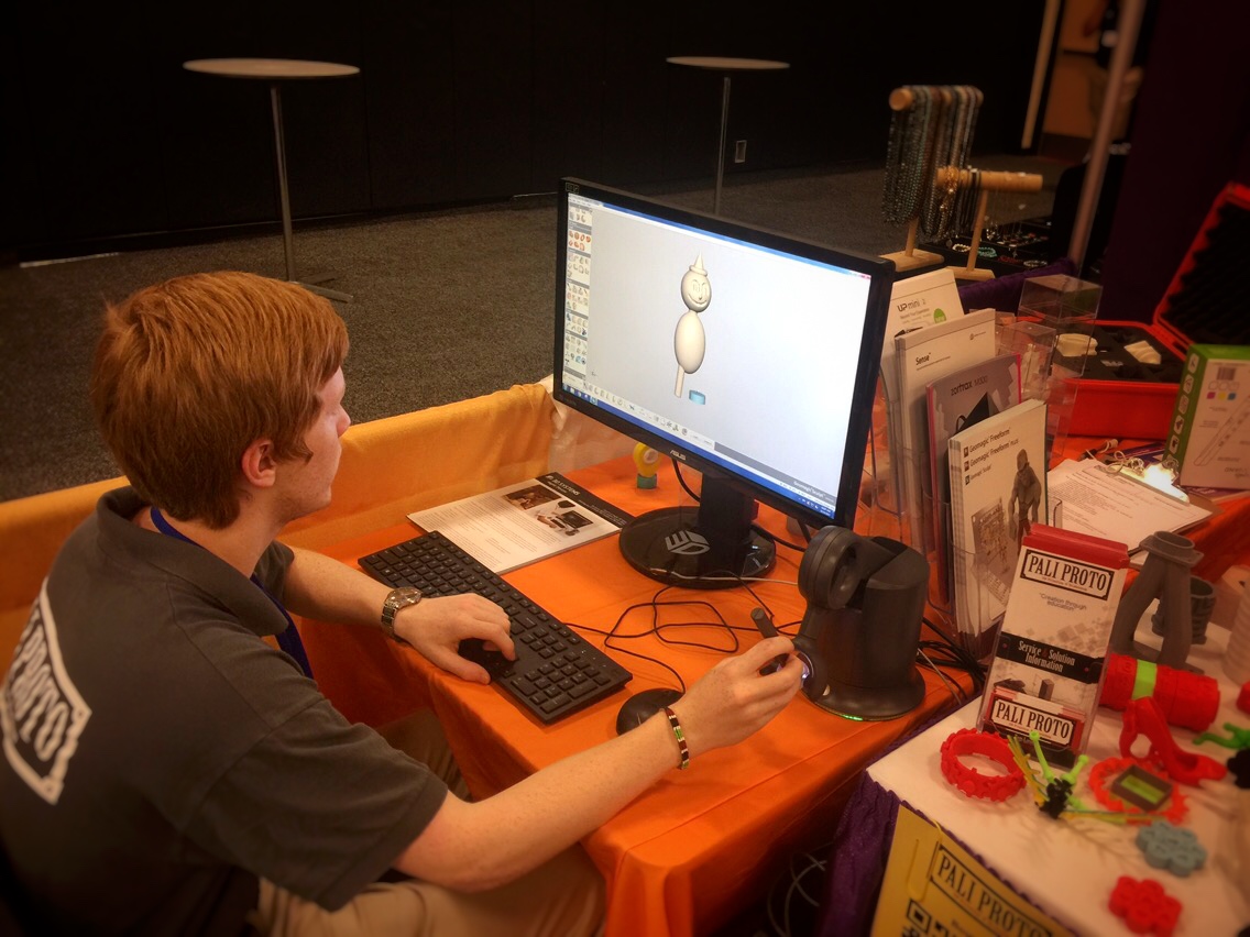 A photo of one of our team members sitting at a computer using a 3D Systems Haptics tool. On the screen is a 3D model that he is manipulating. Various collateral and media are strewn across the table.